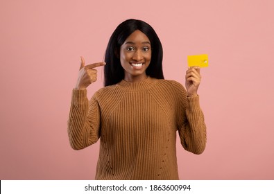 Easy shopping concept. Happy black woman pointing at credit card and smiling on pink studio background. Gorgeous African American lady promoting bank services, advertising online payments