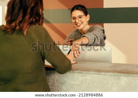 Easygoing receptionist assisting a woman with signing in to an office. Friendly receptionist showing a woman where to sign on a clipboard. Young woman working at the front desk of a convention centre.