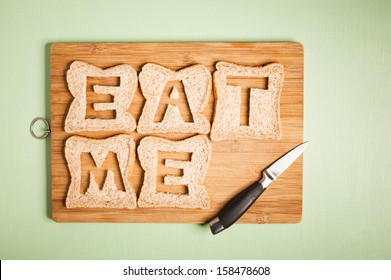 Eat me text carved out of brown bread slices on wooden chopping board with chef's kitchen knife, hunger, hungry