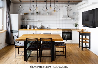 An eat-in kitchen interior design in modern scandinavian style with big wooden table and chairs against light wood floor, bright white walls and furnitures with TV, appliances and hanging light bulbs - Shutterstock ID 1714691389