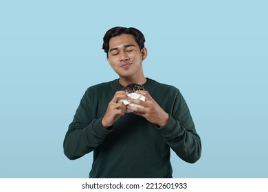 Eating Concept. Asian Man Eating Burger Posing Standing Over Pastel Blue Studio Background. Black Guy Tasting Black Burger Enjoying Unhealthy Junk Food. Nutrition And Overeating Habit