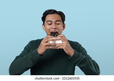 Eating Concept. Asian Man Eating Burger Posing Standing Over Pastel Blue Studio Background. Black Guy Tasting Black Burger Enjoying Unhealthy Junk Food. Nutrition And Overeating Habit