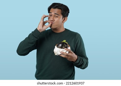 Eating Concept. Asian Man Eating Burger Posing Standing Over Pastel Blue Studio Background. Black Guy Tasting Black Burger Enjoying Unhealthy Junk Food. Nutrition And Overeating Habit