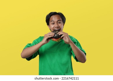 Eating Concept. Asian Man Eating Burger Posing Standing Over Yellow Studio Background. Tasting Black Burger Enjoying Unhealthy Junk Food. Nutrition And Overeating Habit