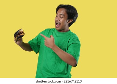 Eating Concept. Asian Man Eating Burger Posing Standing Over Yellow Studio Background. Tasting Black Burger Enjoying Unhealthy Junk Food. Nutrition And Overeating Habit