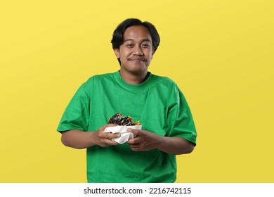 Eating Concept. Asian Man Eating Burger Posing Standing Over Yellow Studio Background. Tasting Black Burger Enjoying Unhealthy Junk Food. Nutrition And Overeating Habit