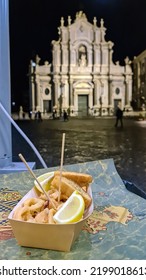 Eating Fried Calamaris With Lemons At Night On The Main Square Of Catania In Sicily, Italy, Europe. View On The Cathedral Of Saint Agatha. Fast Food Tourism. Having Dinner