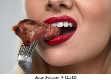 Eating Meat. Closeup Of Woman Mouth With Red Lips, White Teeth Biting Tasty Beef Steak On Fork. Close-up Of Beautiful Female Mouth Eating Delicious Grilled Meat. Nutrition Concept. High Resolution