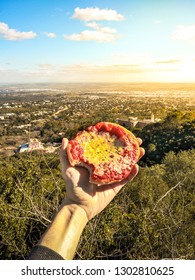 Eating Pizza In Every Place. Pizza Lover: First Person View Of A Hand Holding A Mini Pizza In A Natural Place With A City In The Background At Sunset.