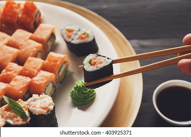 Eating Sushi Rolls. Japanese Food Restaurant, Maki Gunkan Plate Or Platter Set. Closeup Of Hand With Chopsticks Taking Roll. Ginger, Soy, Wasabi Dish At Black Rustic Wood Background.