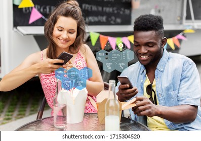 Eating, Technology And People Concept - Happy Mixed Race Couple With Smartphones And Takeaway Chinese Wok And Drinks At Food Truck Over Nutritional Value Chart