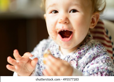 Eating Toddler, One-year-old Child With Messy Mouth And Face Eats Sweets And Laughs. Happy Cute Baby Girl Screams With Delight. Portrait Of Little Kid, Adorable Toddler Having Fun And Food In Kitchen