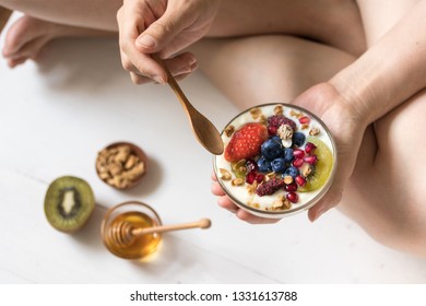 Eating yogurt with strawberry, blueberries, kiwi, granola, pomegranate in a glass bowl and honey on white wooden texture, healthy food and plant-based food concept - Powered by Shutterstock