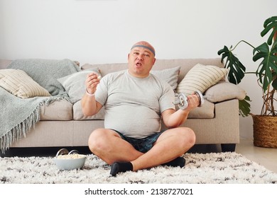 Eats and does exercises. Funny overweight man in casual clothes is indoors at home.