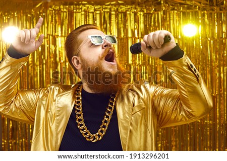 Eccentric singer performing at rock party. Talented man with beard and mustache in extravagant shiny outfit holding microphone and singing songs on stage with gold background at popular music concert