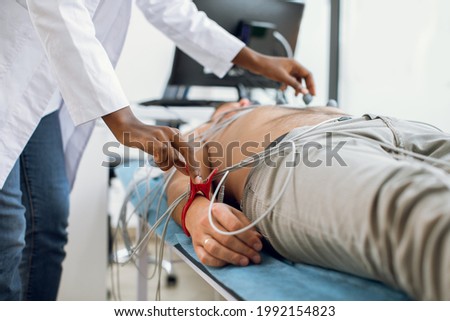 ECG electrocardiography and regular health check concept. Electrocardiogram for heart and pulse measurement in clinic. Female afro nurse sticking vacuum electrodes on patient chest