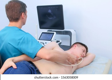 Echocardiography. Man doctor examining guy patient's heart by using ultrasound equipment. He runs ultrasound sensor over man's chest, working on scanner panel, looking at screen.