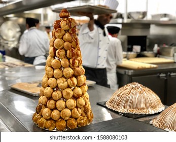 Eclair Pyramid Is On The Table In The Galley Of The Cruise Ship. Chef On A Background Is Walking In The Kitchen.