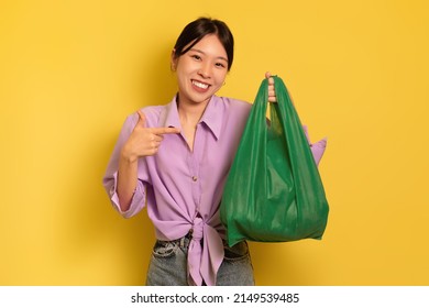 Eco Friendfly Lifestyle. Portrait Of Cheerful Young Asian Woman Pointing At Green Textile Shopping Bag With Products On Yellow Studio Background. Say No To Plastic Concept