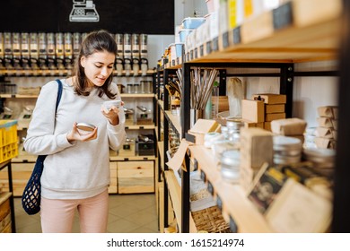 Eco friendly vegan woman customer chooses and buys natural tooth-powder or cream near rack with products in zero waste shop. Eco shopping at local business concept.