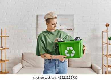 eco-conscious lifestyle, young and tattooed woman with trendy hairstyle holding green recycling box with clothing, sustainable living and environmentally friendly habits concept - Powered by Shutterstock