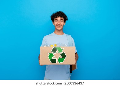 Eco-friendly curly boy guy high school male pupil student teenager with dental braces holding box full of plastic bottles for recycling isolated on blue background copy space