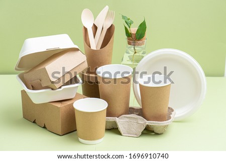 Eco-friendly disposable bamboo cutlery, paper and sugarcane containers for food and drinks over green background. Side view on composition. Plant branch in bottle behind.