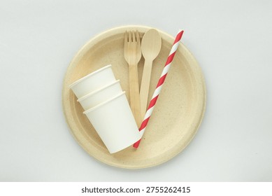 Eco-friendly disposable tableware set with biodegradable plate, wooden fork and spoon, paper cups, and striped paper straw on minimal background. Concept of sustainability, zero waste lifestyle, and plastic free food packaging.