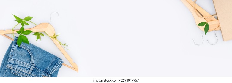 Eco-friendly shopping and slow fashion concept. Upcycling clothes. Jeans, wooden hangers with green leaves, paper shopping bag over light gray background with copy space for text. Banner - Shutterstock ID 2193947957