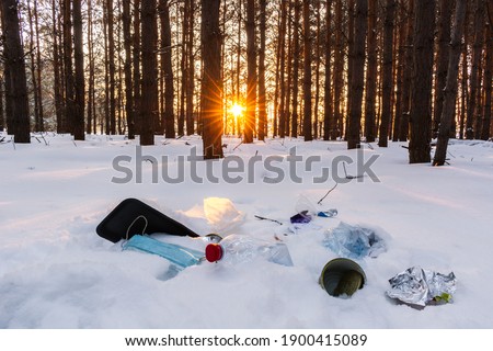 Ecological crisis. Different garbage and trash on snow at beautiful winter forest at bright sunrise with sunbeams through trees. Environmental pollution by garbage. Destructive human influence on