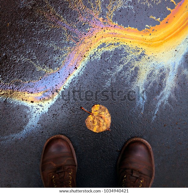 Ecological\
problems. Careless attitude to nature. Environment protection\
concept. Colorful rainbow oil stain on the floor asphalt, yellow\
dead autumn leaf, man\'s shoes. Square 1:1\
shot