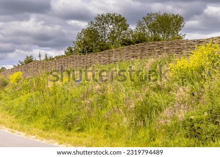 Ecological sound barrier earth wall with wicker fence of willow branches. Flowers en trees are growing on the dike with vegetation. The Netherlands.
