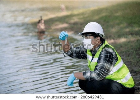 Ecologist analyzing water quality with flask near waterbody wearing protective gear and reflective vest