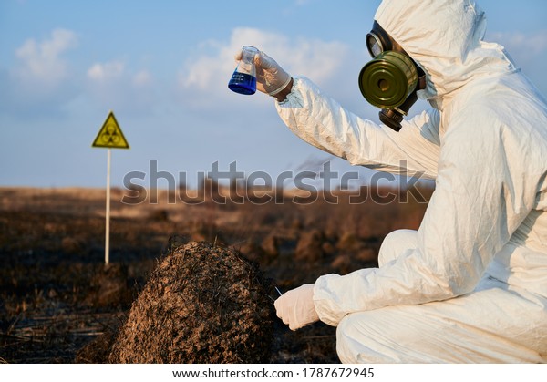 Ecologist in suit, gas mask holding test tube with\
blue liquid while studying burnt grass and soil on scorched\
territory with biohazard sign. Environmentalist doing laboratory\
test in field after\
fire