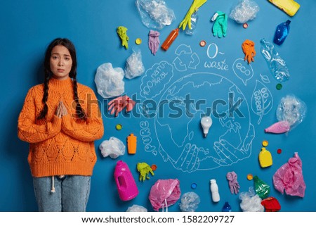 Ecology and land conservation concept. Sad Asian woman stands in praying pose, surrounded with plastic waste, begs for help in cleaning Earth, dressed casually, stands against blue background