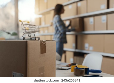 E-commerce business owner moving delivery boxes in the warehouse, small shopping cart in the foreground: online shopping and delivery concept