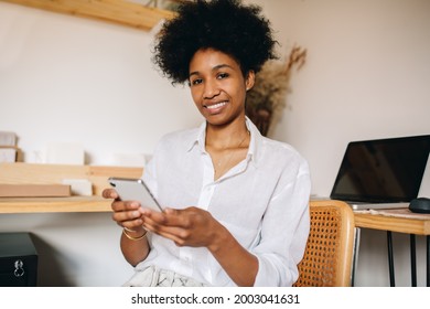 E-commerce Business Owner Smiling At Camera And Checking Customers Reviews For Jewelry Designs. Happy Jewelry Designer Woman Using Phone In Studio.