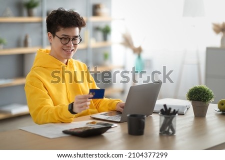 E-Commerce Concept. Portrait Of Cheerful Asian Guy In Glasses Holding Credit Card Shopping Online Using Laptop Sitting At Table At Home. Happy Customer Looking At Bankcard, Selective Focus