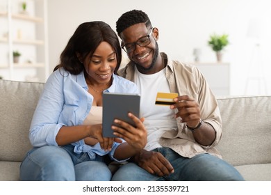 Ecommerce And Easy Payment. Happy Millennial African American Couple Sitting On Couch At Home Using Digital Tablet Choosing What To Buy Looking At Screen, Excited Guy Holding Debit Credit Card