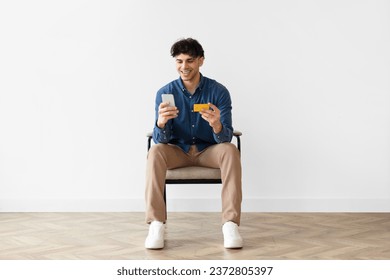 E-Commerce Offer. Contented Middle Eastern Buyer Man Using Credit Card And Phone Shopping Online Sitting In Chair Over White Wall Indoor, Mobile Banking, Payment Application