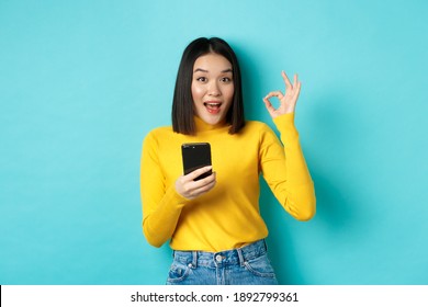 E-commerce And Online Shopping Concept. Portrait Of Asian Woman Showing OK Sign And Using Mobile Phone, Recommend Application, Standing Over Blue Background