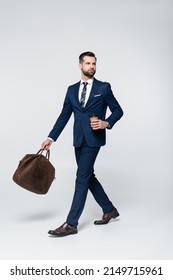 economist in blue suit walking with coffee to go and leather briefcase on grey