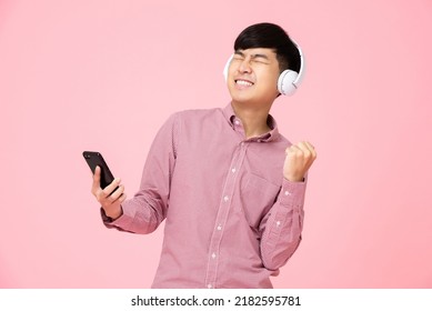 Ecstatic Asian Man Wearing Headphones Listening To Online Streaming Music From Mobile Phone With His Fist Up In Studio Pink Background