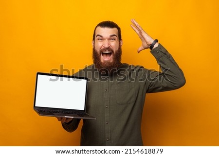 Ecstatic bearded man holding a laptop with blank screen is mind blown. Studio shot over yellow background.