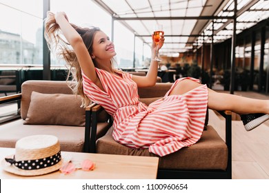Ecstatic blonde girl playing with her hair while chilling in cafe. Inspired young woman in striped dress enjoying champagne in restaurant in summer weekend.