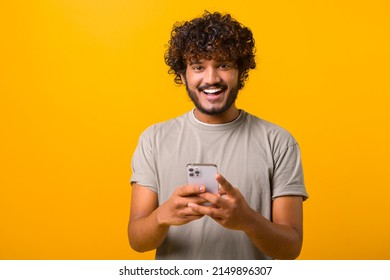 Ecstatic Curly Young Indian Man Standing Isolated On Yellow And Using Trendy Smartphone, Looking At Camera And Smiles Joyfully, Hispanic Millennial Guy Enjoy New Mobile Application Features