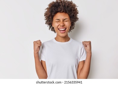 Ecstatic enthusiastic woman with curly bushy hair clenches fists and celebrates success laughs happily dressed in casual basic t shirt isolated over white background. People and triumph concept