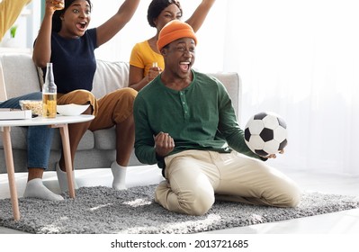 Ecstatic football fans young african american men and women watching game together at home, celebrating victory of their team, raising hands up and screaming, emotional black guy with soccer ball
