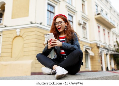 Ecstatic  ginger woman with red lips   chatting by smartphone. Wearing beret and leather jacket sitting on bridge in city center.  - Shutterstock ID 1577649496