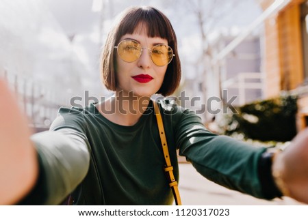 Ecstatic girl in yellow sunglasses making selfie with serious face expression. Outdoor shot of pleased brunette woman in green sweater taking picture on city background.